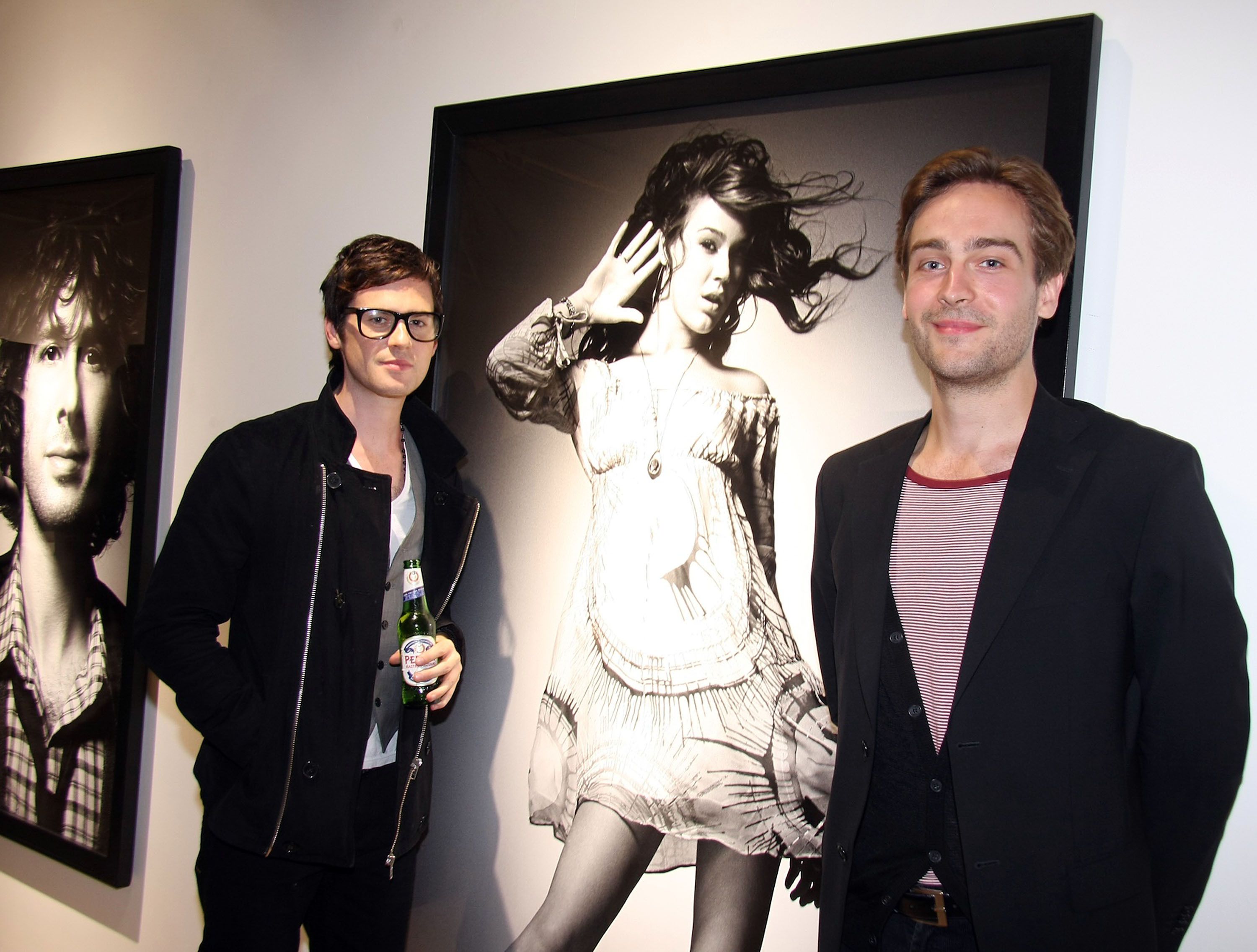 Tom Mison and Tom Riley pose in front of a large, wall-mounted photograph of a wind-blown woman in wearing a dress