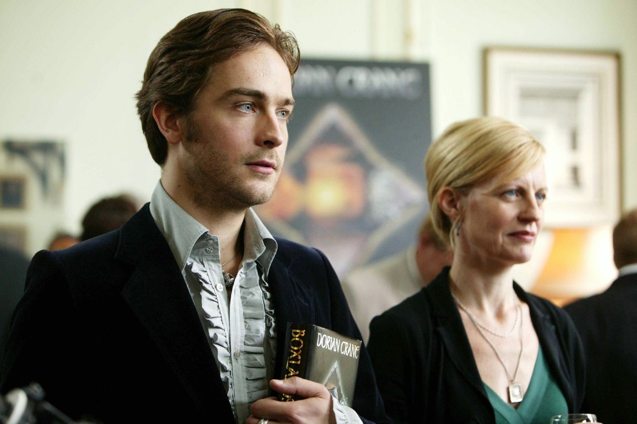 Tom Mison in the tv show Inspector Lewis: Allegory of Love stands in a velvet jacket holding a book with a blond woman next to him